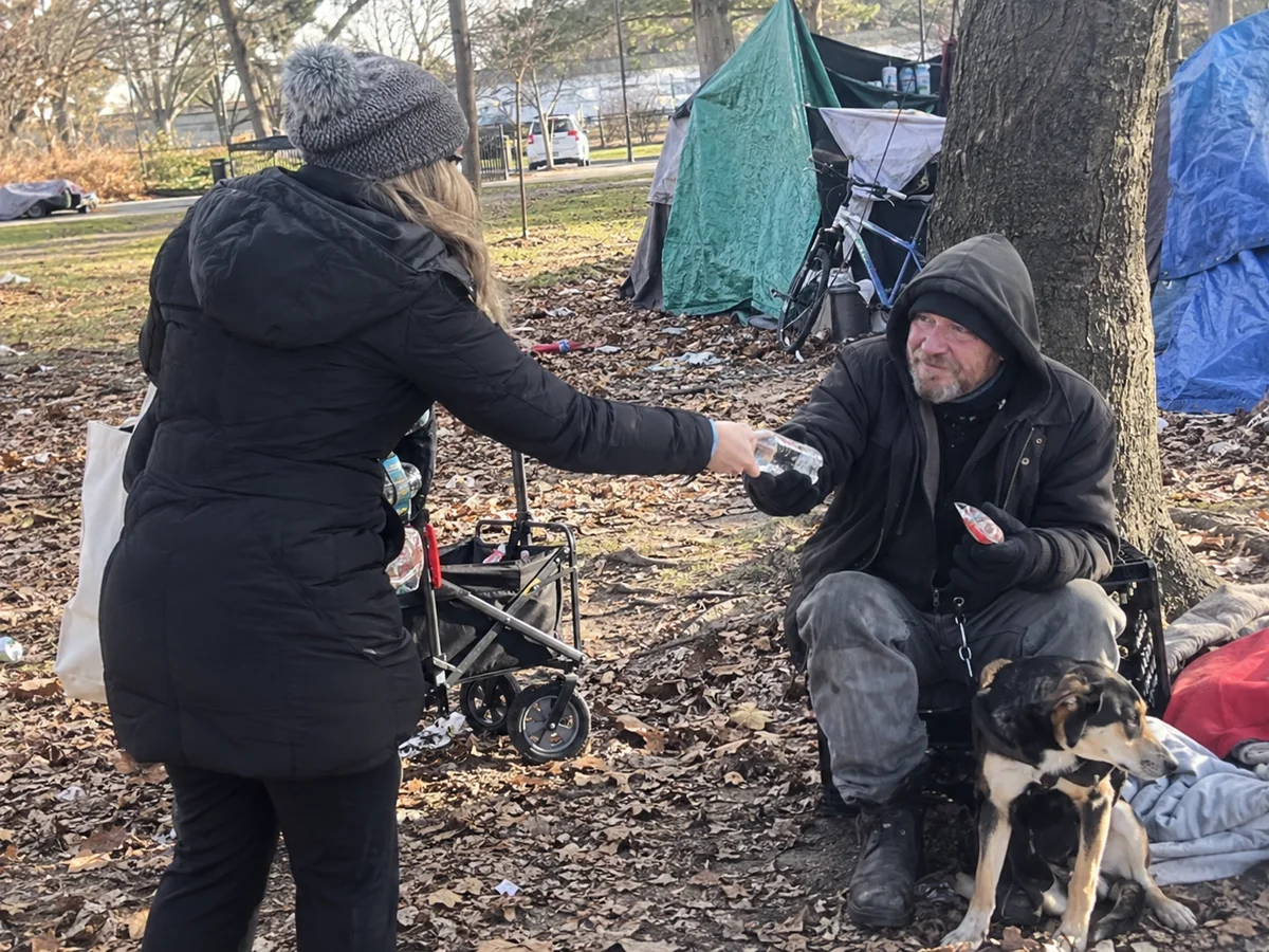 An outreach volunteer handing bottled water to a person beside an encampment.